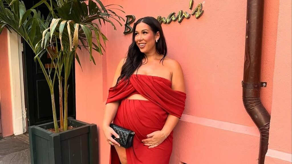 Smiling pregnant woman in a red draped off-shoulder top and gathered high-slit maxi skirt standing against a coral pink wall, holding a black quilted clutch with one hand resting on her baby bump.