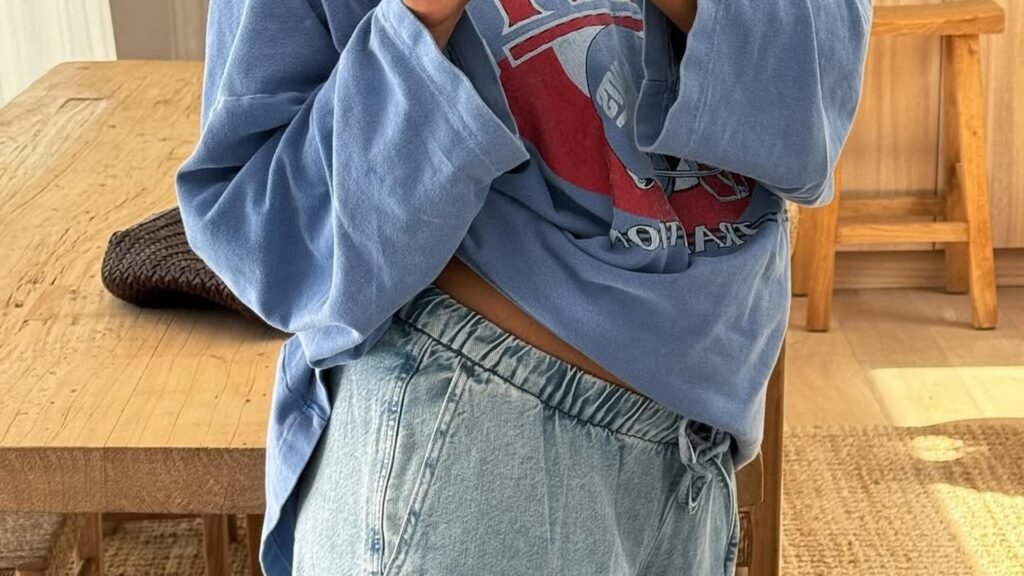 Pregnant woman in an oversized blue graphic sweatshirt and light wash elastic-waist wide-leg jeans, bump visible at the waist gap, styled with regular non-maternity clothes for winter