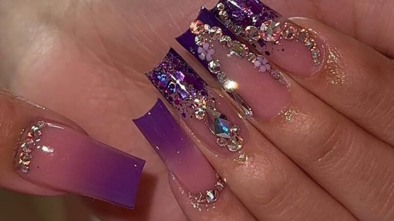 Purple ombre coffin nails with heavy rhinestone and crystal placement, a 3D flower accent, and a teardrop gem pendant, styled as a prom-ready nail look