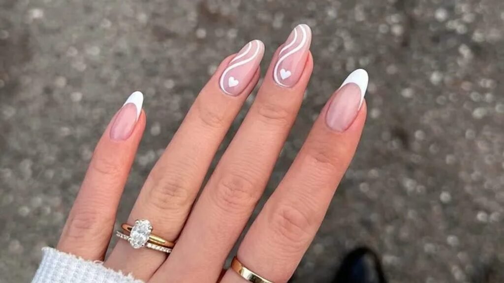 Hand with long almond nails in sheer blush, two with white swirl lines and tiny white hearts and two with classic white French tips, wearing an oval diamond engagement ring stacked with a thin band and a plain gold band.