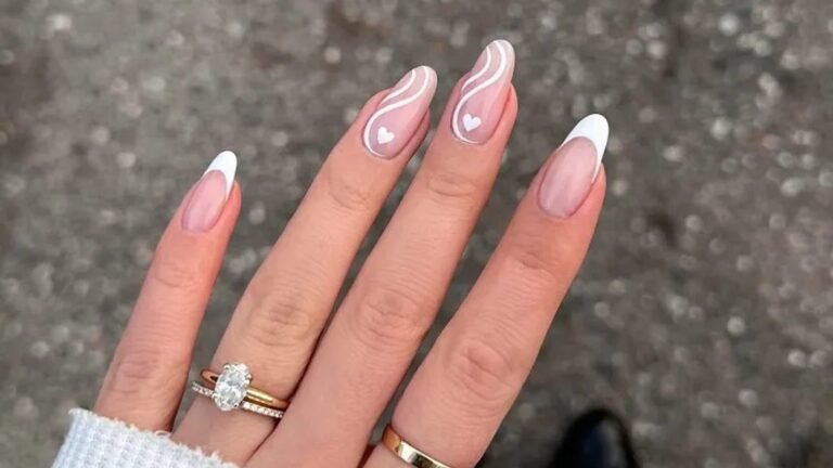 Hand with long almond nails in sheer blush, two with white swirl lines and tiny white hearts and two with classic white French tips, wearing an oval diamond engagement ring stacked with a thin band and a plain gold band.
