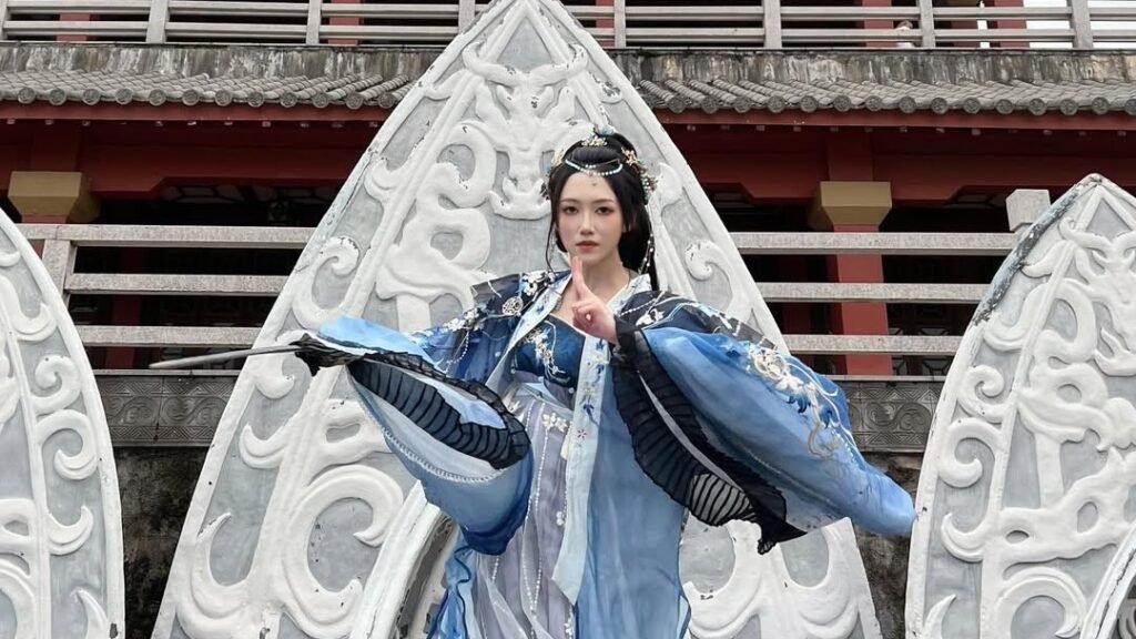 Woman in blue and silver hanfu with flared flowing sleeves posing mid-motion against carved stone archways in front of a traditional Chinese pagoda