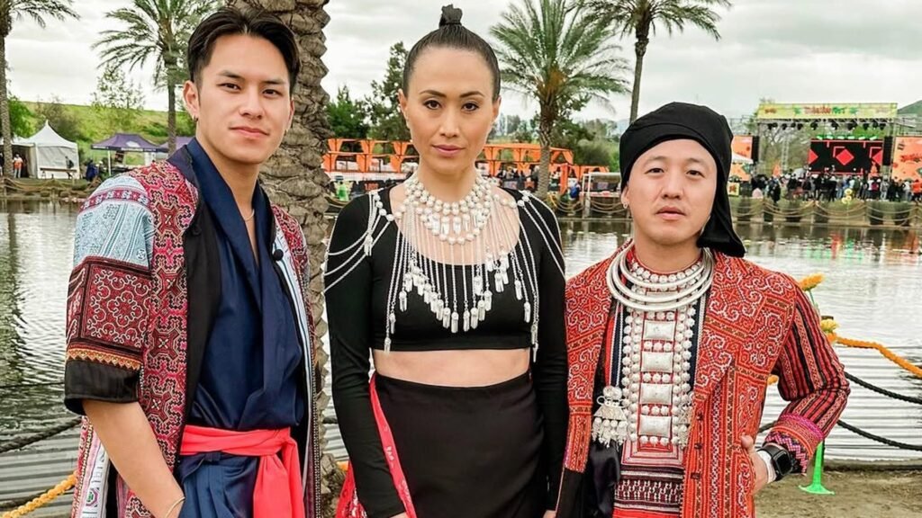 Three people in embroidered red and pink festival coats, a navy outfit with a coral sash, and a black crop top with layered silver necklaces, standing by a lake with palm trees and a festival stage behind them.