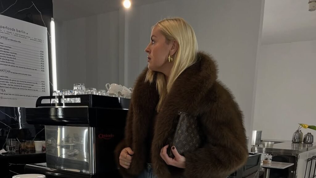 Woman in a chocolate brown fur jacket, wide light-wash jeans, and black pointed boots holding a monogram pouch in a marble café