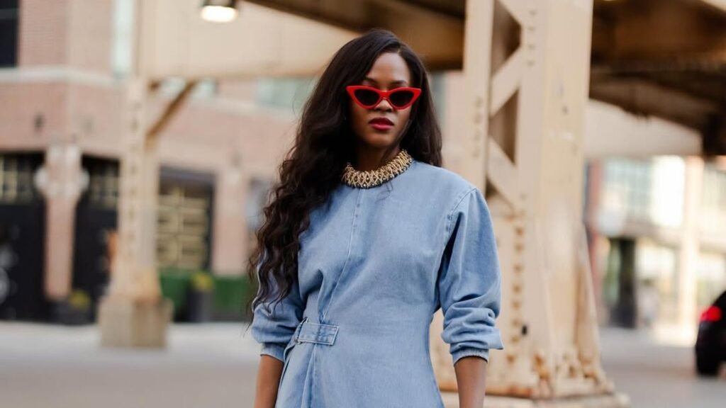 Woman with long wavy dark hair wearing red cat-eye sunglasses, red lipstick, a gold collar necklace, and a light wash denim dress with long sleeves and a belted waist, standing beside a weathered tan steel support column on a city street.