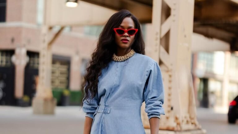 Woman with long wavy dark hair wearing red cat-eye sunglasses, red lipstick, a gold collar necklace, and a light wash denim dress with long sleeves and a belted waist, standing beside a weathered tan steel support column on a city street.