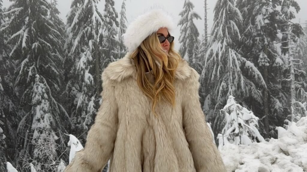 Woman in an oversized cream faux fur coat and matching fur hat standing in front of snow covered pine trees, a cozy one tone winter outfit idea