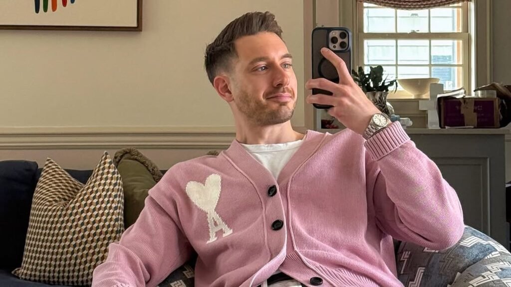 Man in a pink button-up cardigan with a white heart logo over a white tee and white jeans, sitting in a patterned armchair in a cozy living room while taking a mirror selfie.