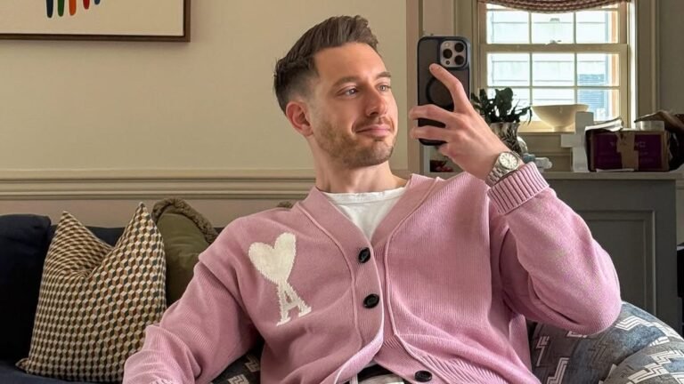 Man in a pink button-up cardigan with a white heart logo over a white tee and white jeans, sitting in a patterned armchair in a cozy living room while taking a mirror selfie.