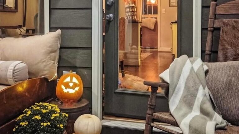 A dark green cottage porch at night with a glowing carved pumpkin, yellow mums, a white pumpkin stack, and an old wood rocking chair draped with a striped throw