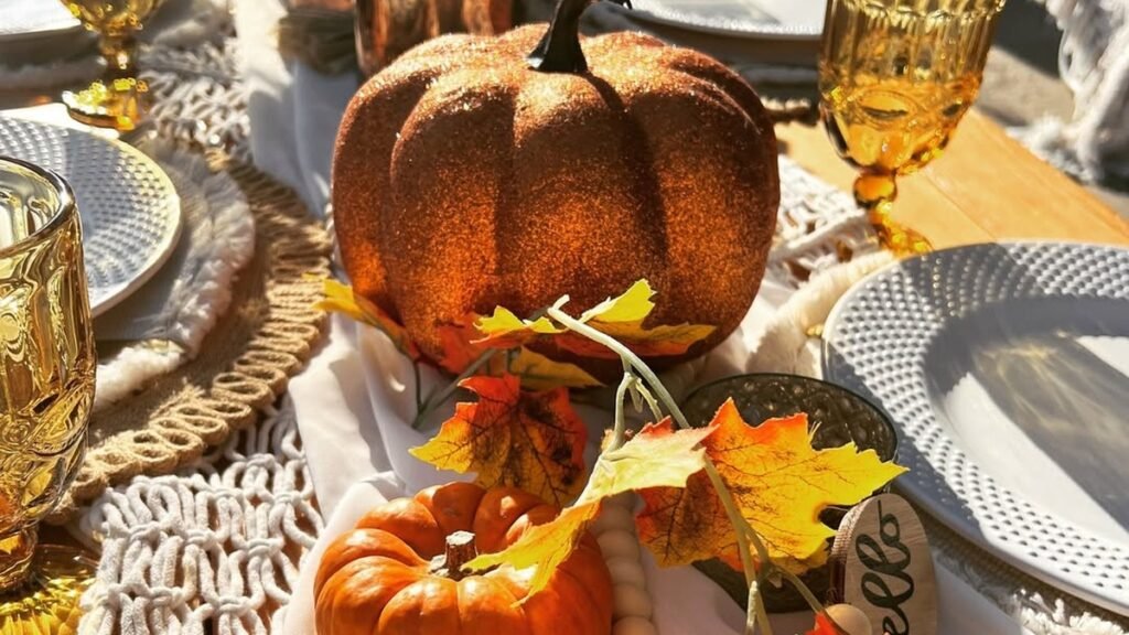 Glittery orange pumpkin centerpiece with autumn maple leaves and amber glassware on an outdoor fall picnic table in golden sunlight