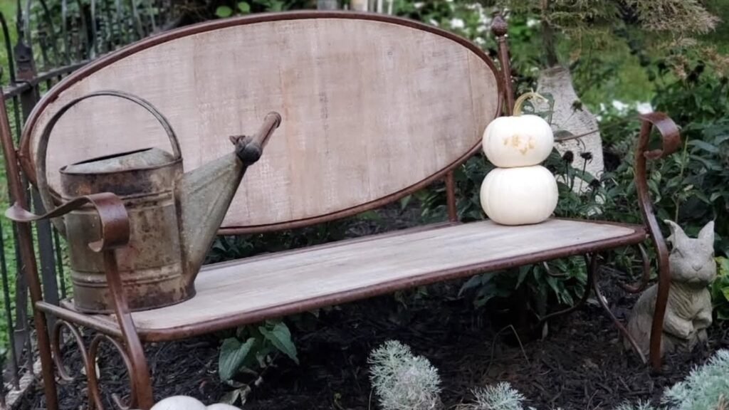 A garden bench styled with white pumpkins, a rustic watering can, and a stone rabbit tucked into a fall garden corner