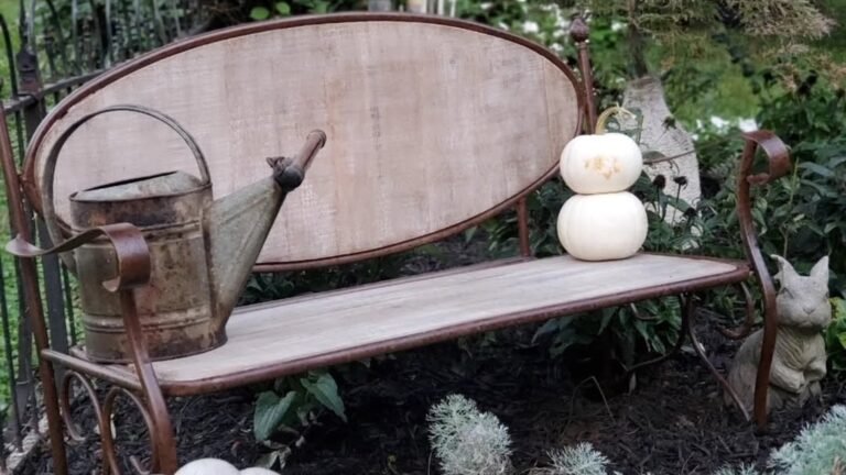 A garden bench styled with white pumpkins, a rustic watering can, and a stone rabbit tucked into a fall garden corner