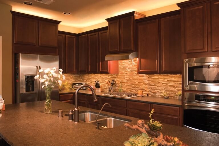 Dark cherry kitchen with warm LED cove lighting above cabinets and glowing backsplash strip lighting below, no ceiling lights on
