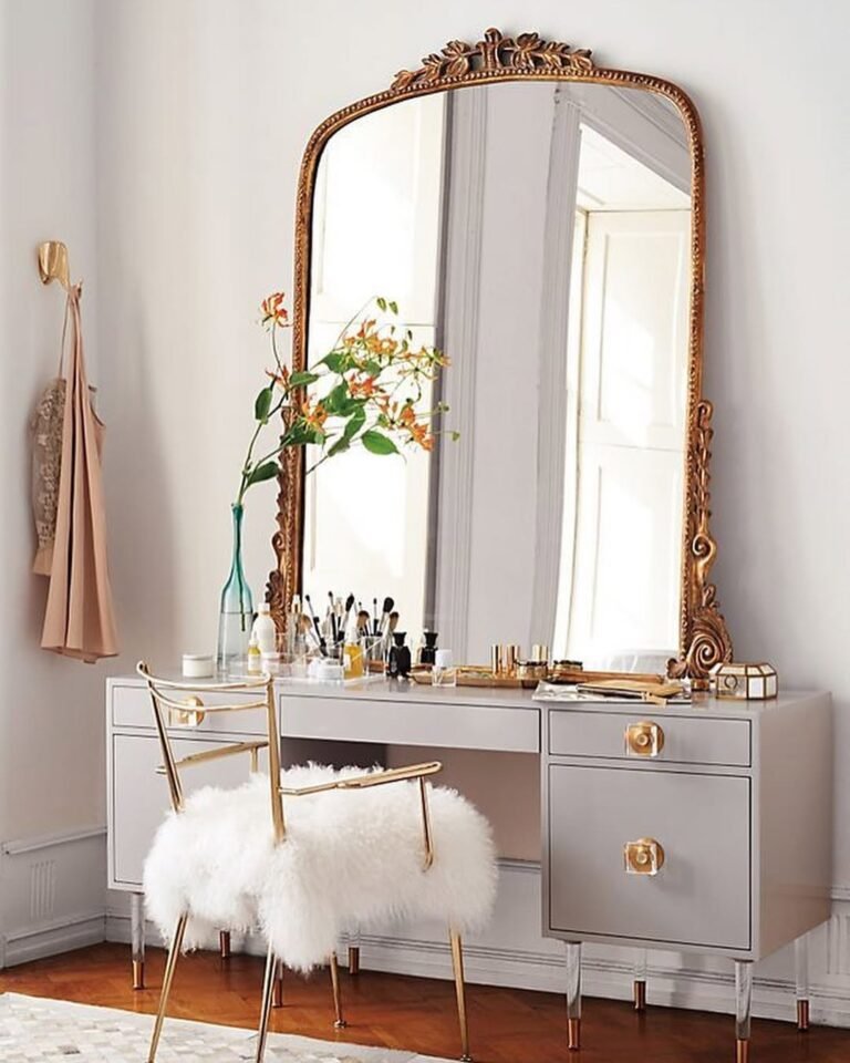 Antique gold arched mirror leaned above a pale gray vanity desk in a bright bedroom corner, styled with a white fur stool, gold-accented chair, and a vase of orange flowers