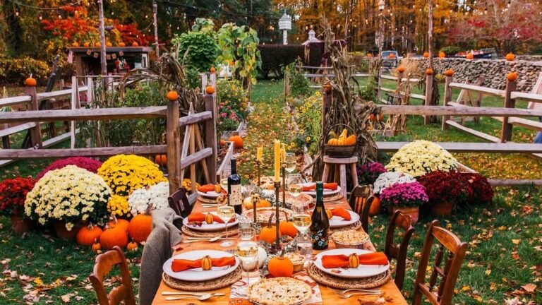 Long outdoor Thanksgiving table set with candles, wine, and a pumpkin pie, flanked by yellow, white, and burgundy mums, with string lights and fiery fall foliage above a wood-fenced garden path