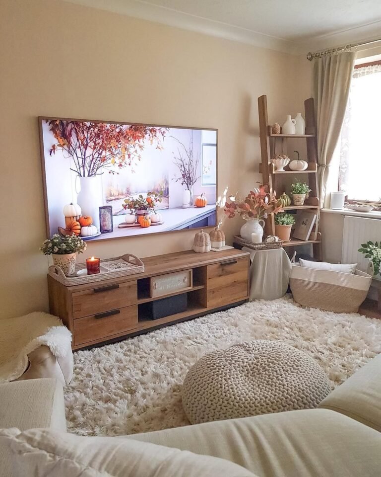 Cozy neutral living room layout ideas with a wood TV console below a wall-mounted TV, cream sofas, a knit pouf on a shaggy rug, and autumn pumpkin decor with a ladder shelf in the corner.
