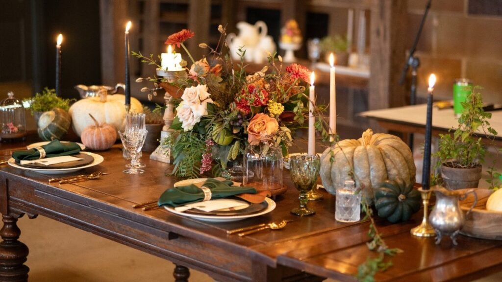 Rustic Thanksgiving decor with heirloom pumpkins, lit taper candles, green napkins, and a harvest floral centerpiece on a dark wood table in a barn
