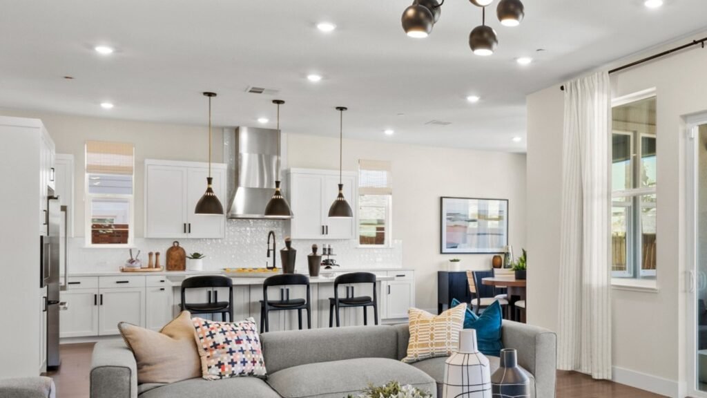 Open concept living room and kitchen with grey sectional sofa, wood coffee table, and white kitchen island in one unbroken sightline under a sputnik chandelier