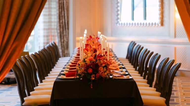 Long Thanksgiving hall table set with lit taper candles and a red and orange floral centerpiece running down the center, chairs lined along both sides