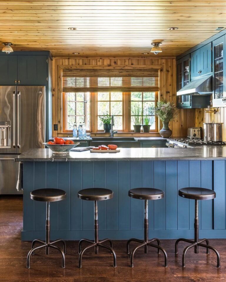 Cabin kitchen with a deep blue painted island and cabinets against raw honey-pine wood walls and ceiling, four bronze bar stools lined up at the island