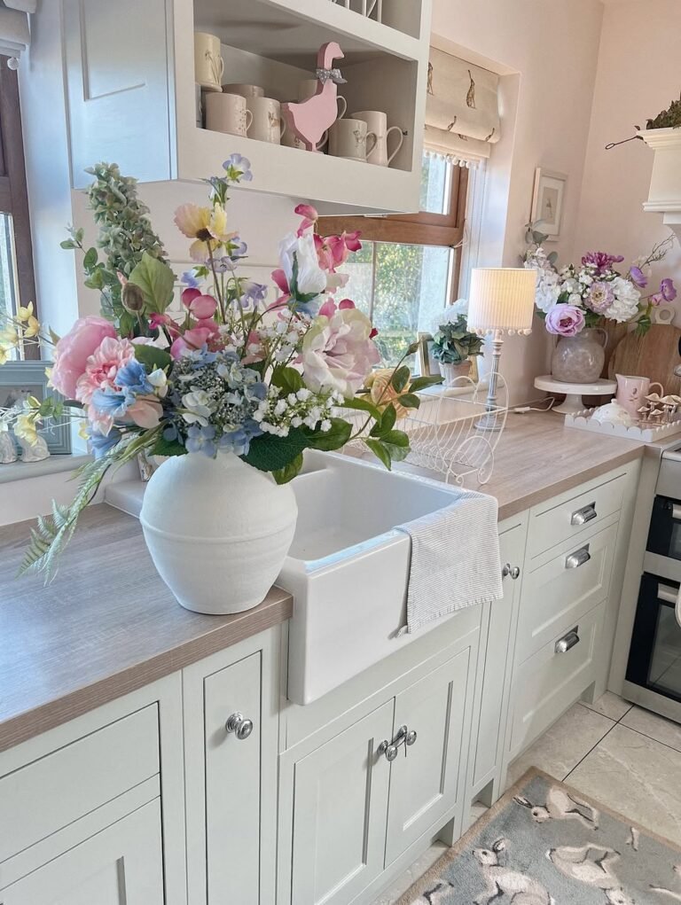 A white farmhouse sink topped with an oversized pastel bouquet of pink and blue flowers in a sage green cottagecore kitchen