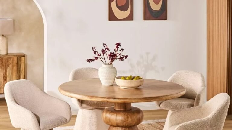 Round wood dining table with sculptural hourglass pedestal base and cream boucle chairs beneath an arched doorway