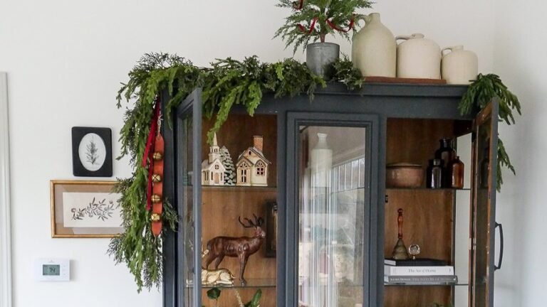 Dark blue glass-front cabinet with open doors revealing shelves styled with mini houses, a deer figurine, amber bottles, and stacked books for layered holiday shelf decor