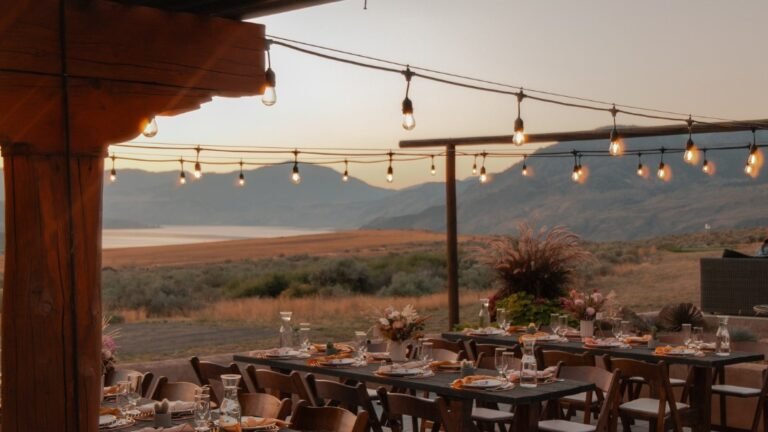 String lights strung across a wooden pergola glow at dusk above a long outdoor dinner table set with plates and flowers, with mountains in the background.