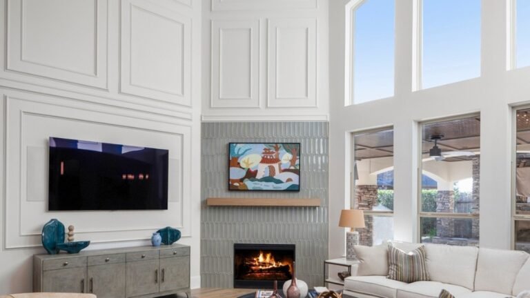 Living room with a plain cream sofa beneath a two-story white paneled wall, arched windows, and a gold ring chandelier