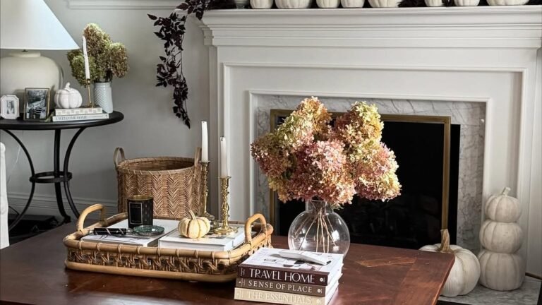 Coffee table styled with a woven tray of books, brass candlesticks, a mini pumpkin, and dried hydrangea in a glass vase, in front of a white fireplace mantel lined with white pumpkins