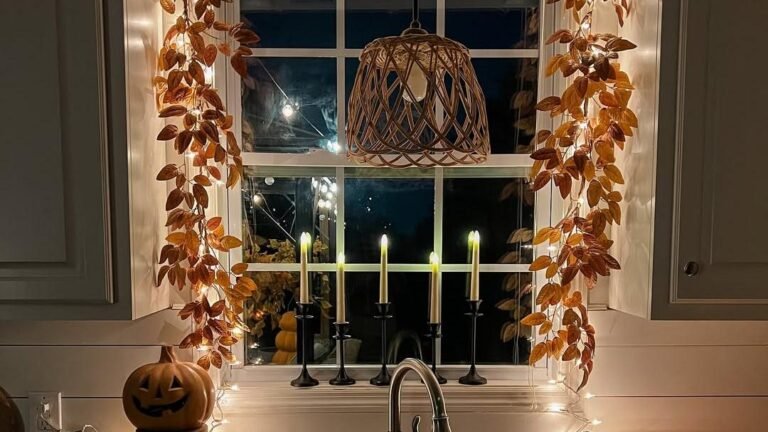 A kitchen window framed in glowing fall leaf garland and string lights, with five lit taper candles lined up on the sill above the sink at night