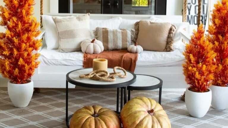 A white hanging porch swing with striped and fringed throw pillows, flanked by two tall orange faux fall trees in white planters, with a black-framed round coffee table holding a lit candle and two large real pumpkins on a patterned rug in the foreground