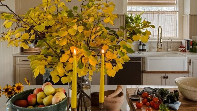 Bright yellow autumn branches and lit taper candles styled on a kitchen island for Thanksgiving decor, with a fruit bowl and sink visible behind