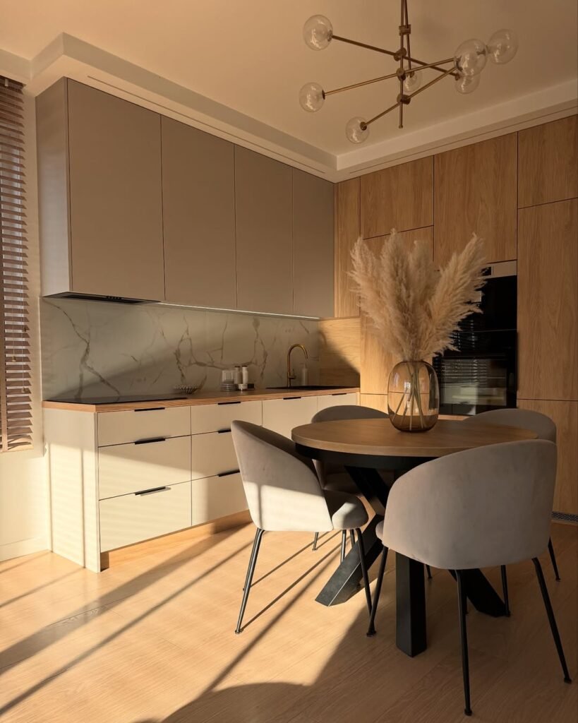 Small apartment kitchen at golden hour with floor-to-ceiling wood and cream cabinets, a slim countertop, and a round dining table tucked into the same compact space.
