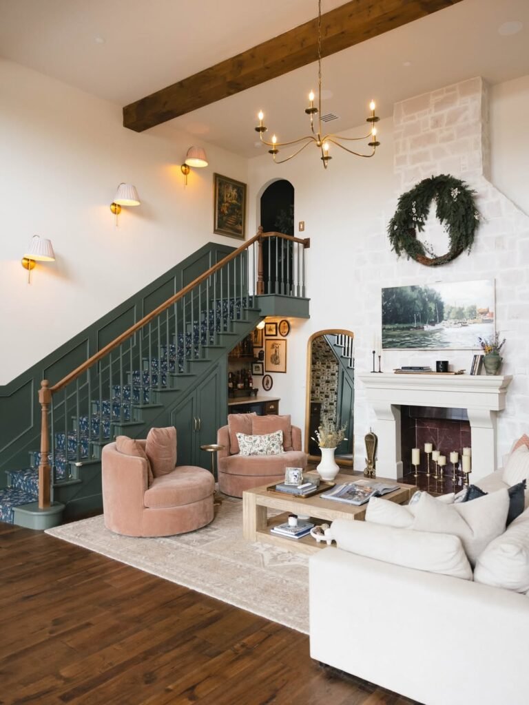 Living room remodel with a forest-green painted staircase and floral runner beside a whitewashed stone fireplace, two blush velvet swivel chairs and a cream sofa around a low oak waterfall table