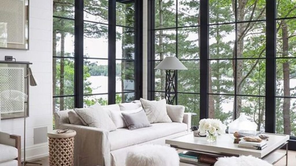 Living room with floor-to-ceiling black-framed cathedral windows overlooking a lake, cream sofa and fur poufs styled to keep the window as the room's focal point