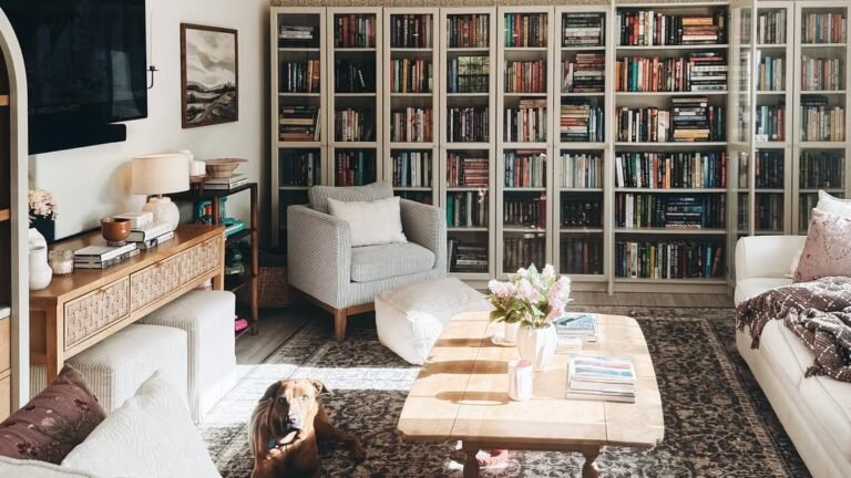 Living room with floor-to-ceiling bookshelves, a striped armchair, and a dog lying on the rug beside a wood coffee table styled with fresh flowers and books