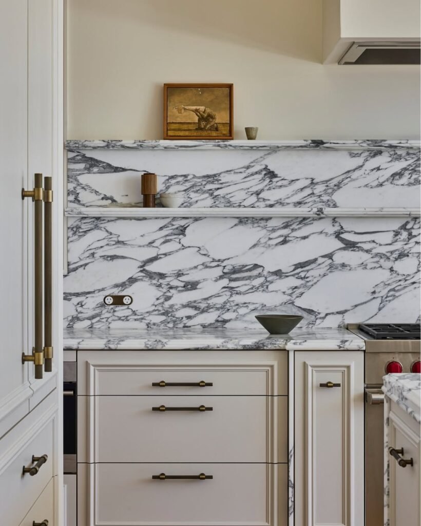 Antique brass cabinet pulls on cream shaker cabinets with a white and gray marble countertop and backsplash