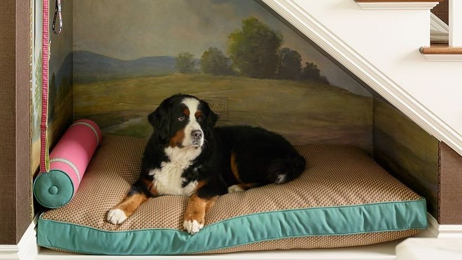 A Bernese mountain dog rests on a plush cushioned dog bed built into a nook under the staircase, with a painted landscape mural backdrop