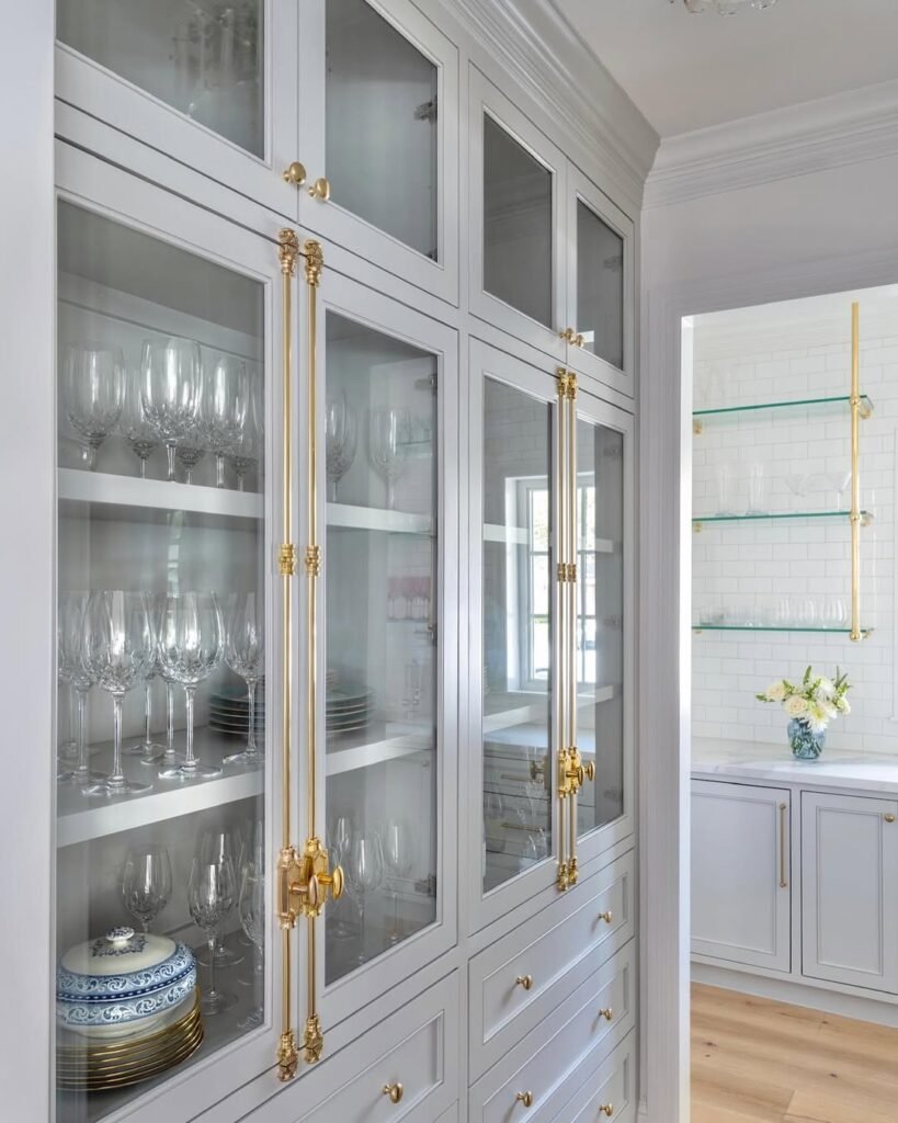 Gray built-in butler's pantry cabinet with glass doors, brass hardware, and crystal stemware displayed on open shelves