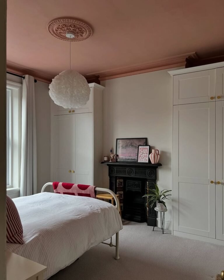 Victorian bedroom with a pink painted ceiling, ornate ceiling medallion, and decorative cornice molding above white wardrobes and a black cast iron fireplace