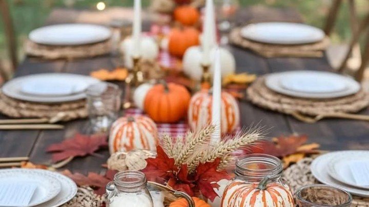 A red and white gingham table runner set with mini pumpkins, milk glass bottles, taper candles, and scattered autumn leaves on a dark wood table, string lights glowing in the background