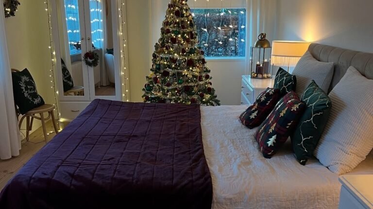 Bedroom with a plum velvet throw on a white quilted bed, tartan and forest-green pillows, a lit Christmas tree by the window, and star string lights hanging in the curtain