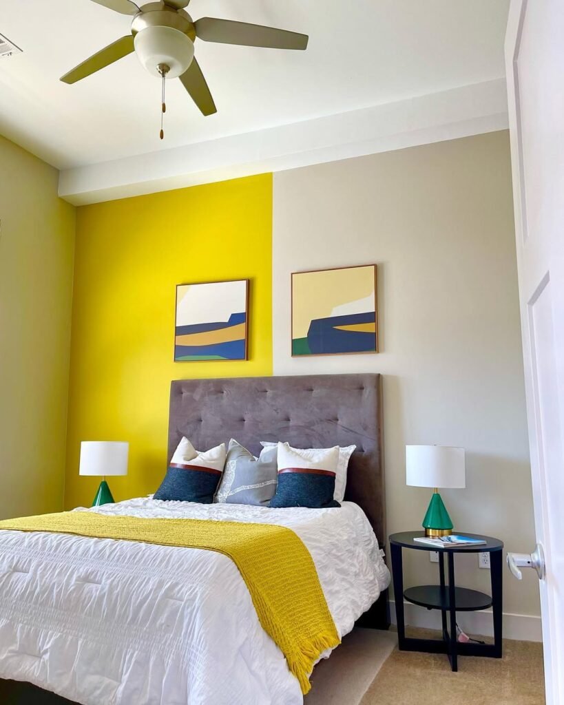 Bedroom with a color-blocked yellow and gray accent wall, gray tufted headboard, matching framed art, and green table lamps