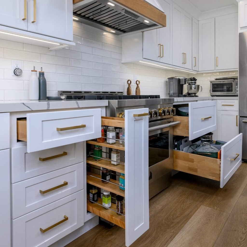 Built-in kitchen storage with three drawers open at once around a pro range, showing a vertical spice rack, deep pot drawer, and wide pan pull-out with white shaker fronts and gold pulls
