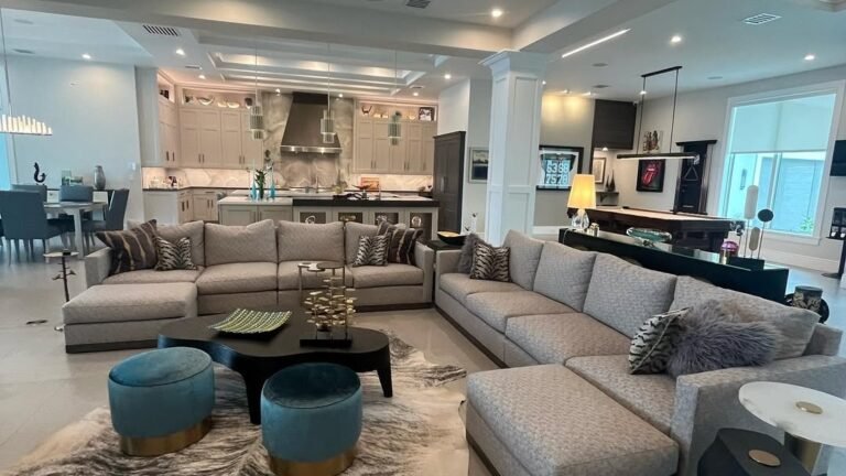 Long living room with two gray sectional sofas facing each other, teal velvet ottomans on a cowhide rug, and an open sightline to a pool table and kitchen beyond.