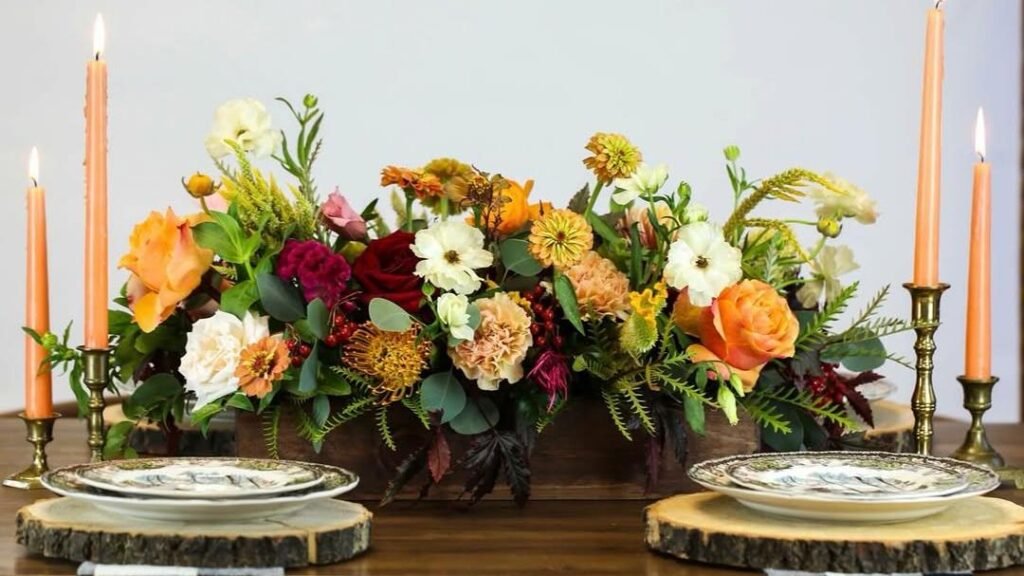 Loose fall floral centerpiece in a wooden box with roses, ranunculus, and greenery on a Thanksgiving table set with vintage plates, wood-slab chargers, and lit taper candles