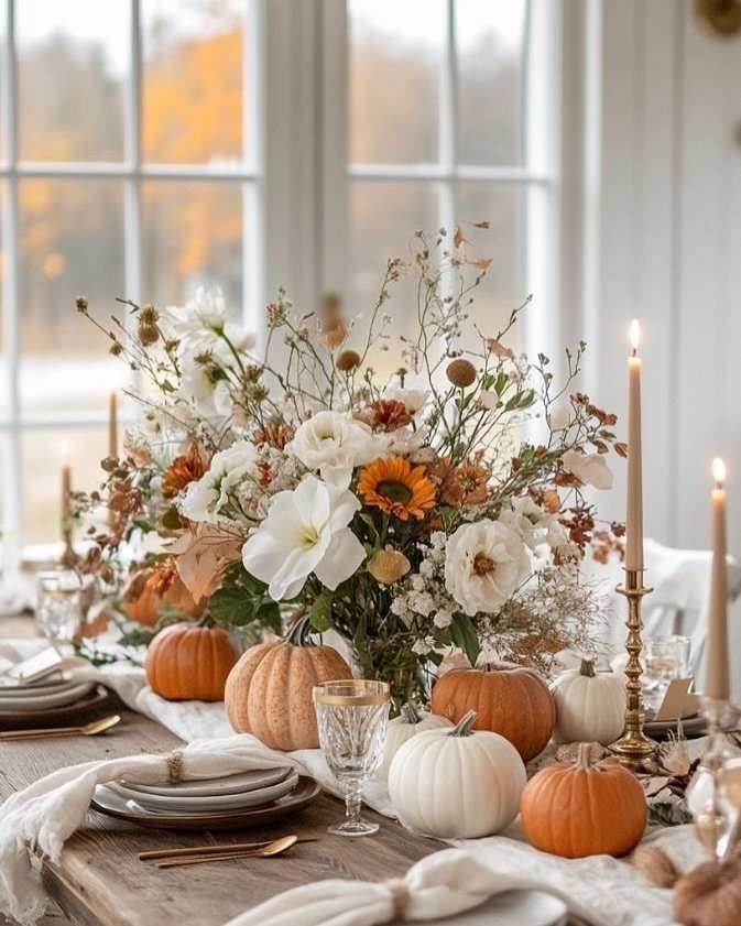 White pumpkin and floral centerpiece with warm-toned flowers, gold candlesticks, and lit taper candles on a rustic Thanksgiving table