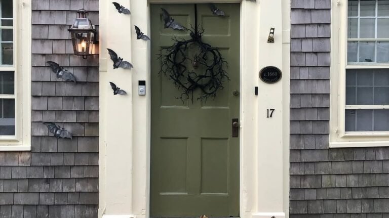 Black front door with scattered bat decorations and a black wreath, flanked by white potted trees and a moon-and-bats doormat on a stone porch